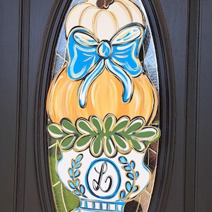 May include: A decorative pumpkin-themed door hanger. The design features a stacked pumpkin with a blue bow, a white pumpkin top, and a vase with the letter "L". The color palette includes orange, white, blue, green, and brown.