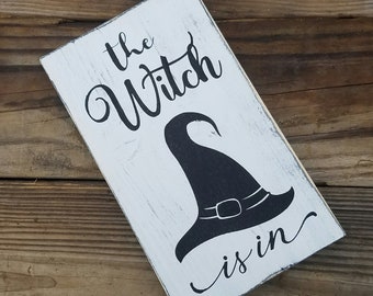 May include: White wooden sign with black text and a black witch's hat silhouette. The text reads "The Witch is in."