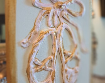 May include: A decorative wall art piece featuring a relief of ballet pointe shoes and a bow. The shoes and bow are painted in a soft pink and accented with gold. The artwork is mounted on a light beige canvas with a gold border.