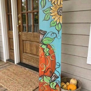 May include: A tall, rectangular wooden porch sign painted with a light blue background. The design features two large orange pumpkins stacked vertically, surrounded by green leaves and vines, with yellow sunflowers at the top and bottom.