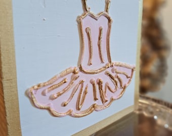 May include: A wooden block with a white painted front featuring a raised relief of a pink and gold ballerina tutu. The edges of the block are painted gold. The tutu design includes straps and a layered skirt.