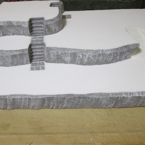 May include: A three-tiered, white, foam-based terrain model with gray stone-like details. The model features a set of stairs and a curved opening.