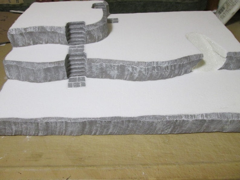 May include: A three-tiered, white, foam-based terrain model with gray stone-like details. The model features a set of stairs and a curved opening.