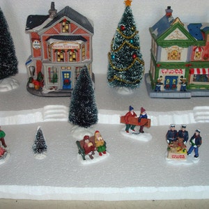 May include: A miniature Christmas village scene with a red brick building with a sign that reads "SALOON GIFT SHOP", a green and white building with a sign that reads "THE TOY SHOP", a Christmas tree, and several miniature figures.