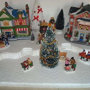 May include: A miniature Christmas village scene with a white snowy base, two houses, a tree, and several figures. The houses are decorated with lights and have signs that read "Toy Shop" and "Walton's Gift Shop".