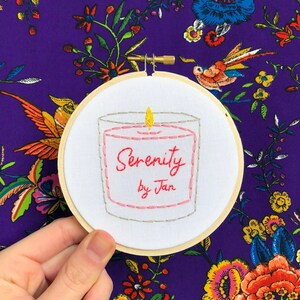 May include: A white embroidery hoop with a hand-stitched design of a candle with a pink and gray body and a yellow flame. The candle has the text "Serenity by Jan" stitched in red and gray thread.