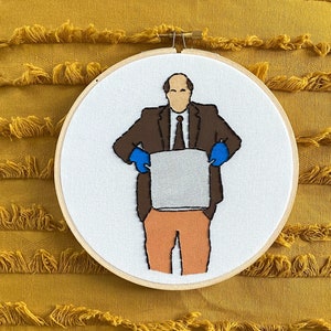 Könnte beinhalten: Stickerei in einem runden Rahmen mit einem Comic-Charakter, der einen braunen Anzug, blaue Handschuhe trägt und eine weiße Box hält. Der Charakter hat braunes Haar und einen Schnurrbart.