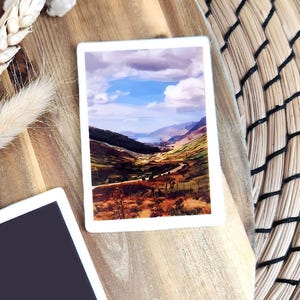 May include: A rectangular print depicting a valley landscape with a winding road. The artwork shows a blue sky with clouds, green and brown hills, and a distant body of water. The print is placed on a wooden surface.
