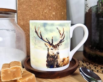 May include: White ceramic mug with a geometric stag illustration. The mug is on a small wooden coaster. Fudge pieces and a glass jar are in the foreground. A corkboard and a glass terrarium are in the background. The mug is ideal for hot beverages.