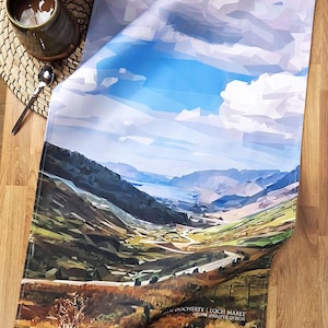 May include: A folded tea towel with a geometric landscape design depicting a blue sky, mountains, and a winding road. The towel has the text "LOCH MAREE" and "LOUISE JENNIFER DESIGN". It is placed on a wooden surface.