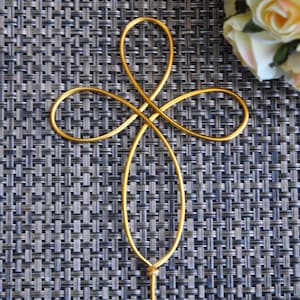 May include: A gold wire cross with a simple, elegant design. The cross is made of a single piece of wire that is bent into a shape that resembles a cross. The wire is smooth and shiny, and the cross is a beautiful piece of jewelry.