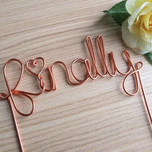 May include: A copper wire cake topper with the word "finally" spelled out in cursive. The "f" has a heart shape in the middle of the letter.