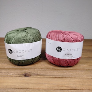 May include: Two balls of crochet thread, one olive green and one dusty rose pink. Each ball features a white paper label with the black text "KP CROCHET" and "Curio" printed on the front. The thread has a smooth, mercerized appearance.