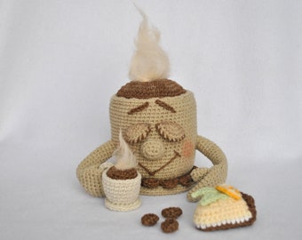 May include: A crocheted coffee cup character with a brown face, holding a smaller crocheted cup. The character is sitting on a white surface with a crocheted slice of cake and brown chocolate candies.