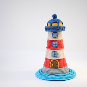 Può includere: Un giocattolo faro in crochet con strisce rosse, bianche e blu, un tetto blu e una finestra gialla. Il faro è in piedi su una base blu che ricorda l'acqua.