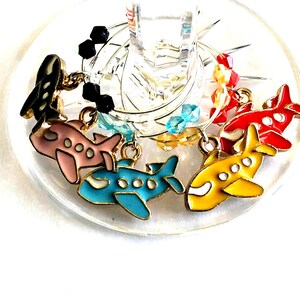 May include: Set of six colorful airplane-shaped wine glass charms. Each charm features a different color airplane with gold accents and crystal beads.