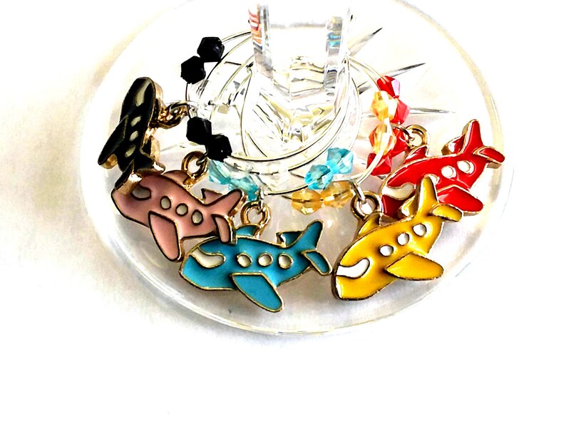 May include: Set of six colorful airplane-shaped wine glass charms. Each charm features a different color airplane with gold accents and crystal beads.