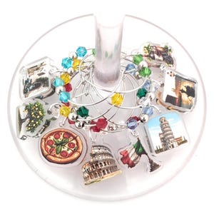 May include: A set of wine glass charms with Italian-themed designs. The charms include images of the Leaning Tower of Pisa, the Colosseum, pizza, and the Italian flag. The charms are attached to silver-colored rings and adorned with colorful beads.