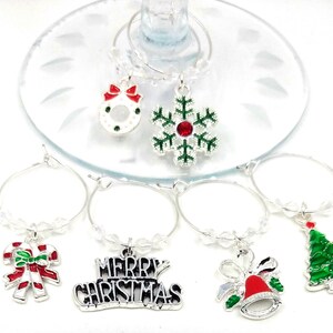 May include: Set of six silver Christmas themed wine glass charms. The charms feature a snowflake, a wreath, a candy cane, a Christmas tree, a bell with a red bow, and a charm that says "Merry Christmas".