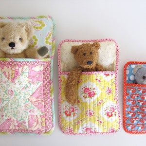 May include: Three quilted fabric sleeping bags for stuffed toys. The sleeping bags are in pink, yellow, and blue with different patterns. Each sleeping bag has a stuffed toy inside.