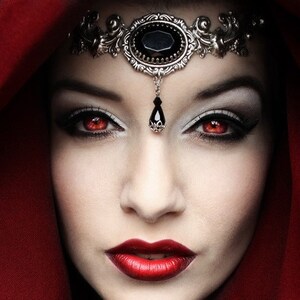 May include: A woman with red lipstick and red eyes wears a red hooded cloak and a silver headband with a black gemstone.