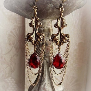 May include: Gold dangle earrings with a red teardrop-shaped crystal and a filigree design.