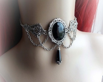 May include: A silver choker necklace with a large black oval gemstone and a smaller black teardrop pendant. The necklace features intricate filigree details and a chain design.