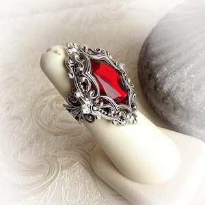 May include: A silver ring with an ornate design and a large, red, faceted gemstone.