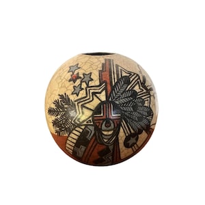May include: A round, cream-colored ceramic vase with a cracked glaze. The vase features a Native American-inspired design with geometric shapes, stars, and feathers in black, red, and gray. The vase has a small opening at the top.