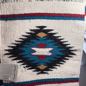 May include: Cream-colored woven textile with a geometric design. The central pattern features a black, teal, red, and gold diamond shape. Red and blue stripes border the top and bottom. The textile has a fringed edge and a tag that reads "APOTEC WEAVING".