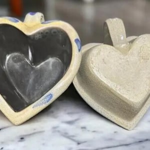 Puede incluir: Dos platos de cerámica en forma de corazón, uno con interior esmaltado y el otro con superficie lisa sin esmaltar. Ambos platos son de color crema con detalles azules.