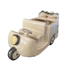 May include: A beige ceramic planter shaped like a vintage three-wheeled vehicle. The planter features a steering wheel, a headlight, and a small cargo area. The wheels are black and the headlight is yellow.