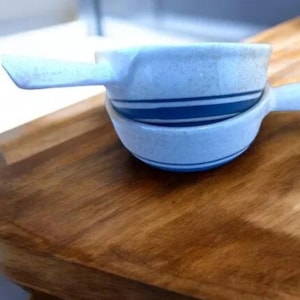 Pode incluir: Duas tigelas de cerâmica branca com listras azuis. As tigelas têm alças e estão empilhadas uma em cima da outra.