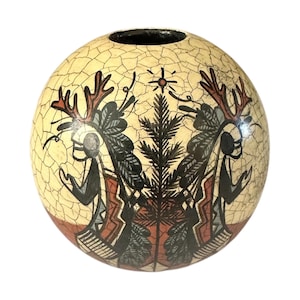 Puede incluir: Un jarrón redondo de cerámica color crema con un aspecto agrietado y envejecido. El jarrón presenta un diseño simétrico de dos figuras con cornamentas y tocados, flanqueando un árbol central. Las figuras son en negro, rojo y gris.