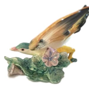 May include: A ceramic figurine of a yellow bird with black and brown wings perched on a green leafy base with a pink flower.