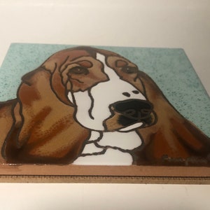 May include: A ceramic tile featuring a hand-painted illustration of a brown and white basset hound dog. The dog's head and shoulders are visible, with a light blue background.