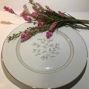 Könnte beinhalten: Ein weißer Teller aus Keramik mit goldfarbenem Rand und einem zarten Blumendesign in Rosa- und Grüntönen. Der Teller ist leer und bereit zum Gebrauch.