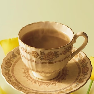 Puede incluir: Un juego de taza y platillo beige con detalles florales y lineales. La taza está llena de un líquido marrón, posiblemente té o café. El juego está colocado sobre una superficie verde claro, con flores amarillas en el fondo.