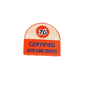 May include: A vintage patch with a white and orange arched top and a rectangular orange bottom. The top features the number "76" in a red circle. The bottom section reads "CERTIFIED AUTO CARE SERVICE" in blue text.