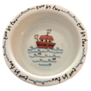 May include: White ceramic bowl with a blue and white border. The bowl features a hand-painted illustration of Noah's Ark with animals on board, sailing on blue waves with fish. The text "two by two" is repeated around the rim of the bowl.