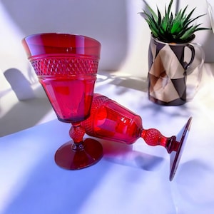 May include: Two red glass goblets with a diamond pattern on the bowl and a textured stem. The goblets are on a white surface.