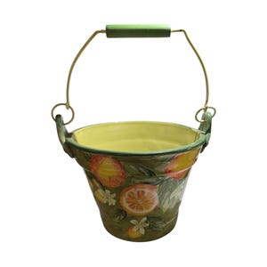 May include: A ceramic bucket with a green handle and a metal wire handle. The bucket is painted with a design of oranges, lemons, and white flowers on a green background. The interior is yellow.