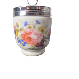 May include: A white porcelain egg coddler featuring a colorful floral pattern with pink roses, yellow blossoms, and blue flowers. The container has a polished silver-tone metal lid with a circular ring handle at the top and a rounded base.