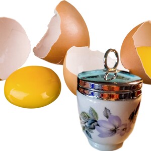 May include: A white porcelain egg coddler with a silver-coloured metal lid and a ring handle, decorated with purple flowers and green leaves, positioned next to three brown eggshell fragments and a single bright yellow raw egg yolk.