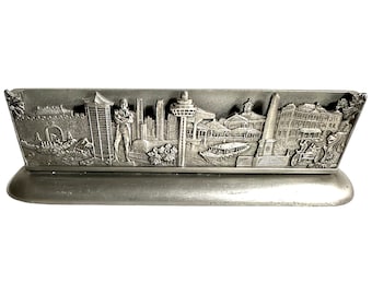 Könnte beinhalten: Ein silberner Metall-Schreibtisch-Organizer mit einem 3D-geprägten Design der Skyline von Singapur. Das Design umfasst ikonische Sehenswürdigkeiten wie das Marina Bay Sands, das Singapore Flyer und den Merlion.