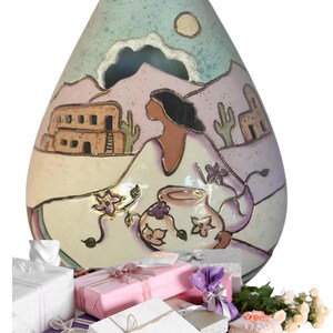 May include: A decorative ceramic vase with a Southwestern design, featuring a woman, buildings, and cacti. The vase is surrounded by wrapped gift boxes in various colors, including white, pink, and purple. A bouquet of white roses is also present.