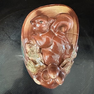May include: A brown ceramic squirrel figurine with a glossy finish. The squirrel is surrounded by leaves and acorns, set against a brown oval background. The figurine has a detailed, three-dimensional design.