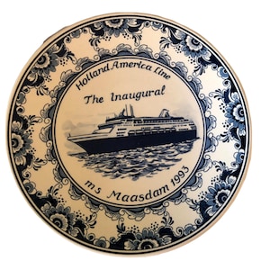 Puede incluir: Un plato de cerámica blanco con un borde floral azul y blanco. El centro del plato presenta una ilustración en blanco y negro de un crucero con el texto "Holland America Line The Inaugural m.s. Maasdam 1993".