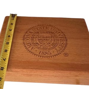 Puede incluir: Una tabla de cortar de madera con el sello de la Universidad Estatal de Arizona grabado en la superficie. La tabla mide aproximadamente 23 cm de largo. La madera tiene un tono cálido y natural.
