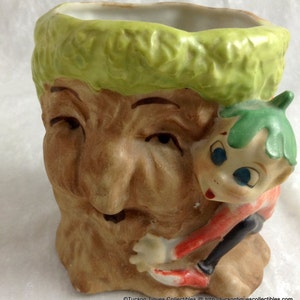 Puede incluir: Una maceta de cerámica con forma de tronco de árbol con una pequeña figura de duende trepando por el lado. El duende tiene el pelo verde, los ojos azules y lleva un atuendo rojo y negro.